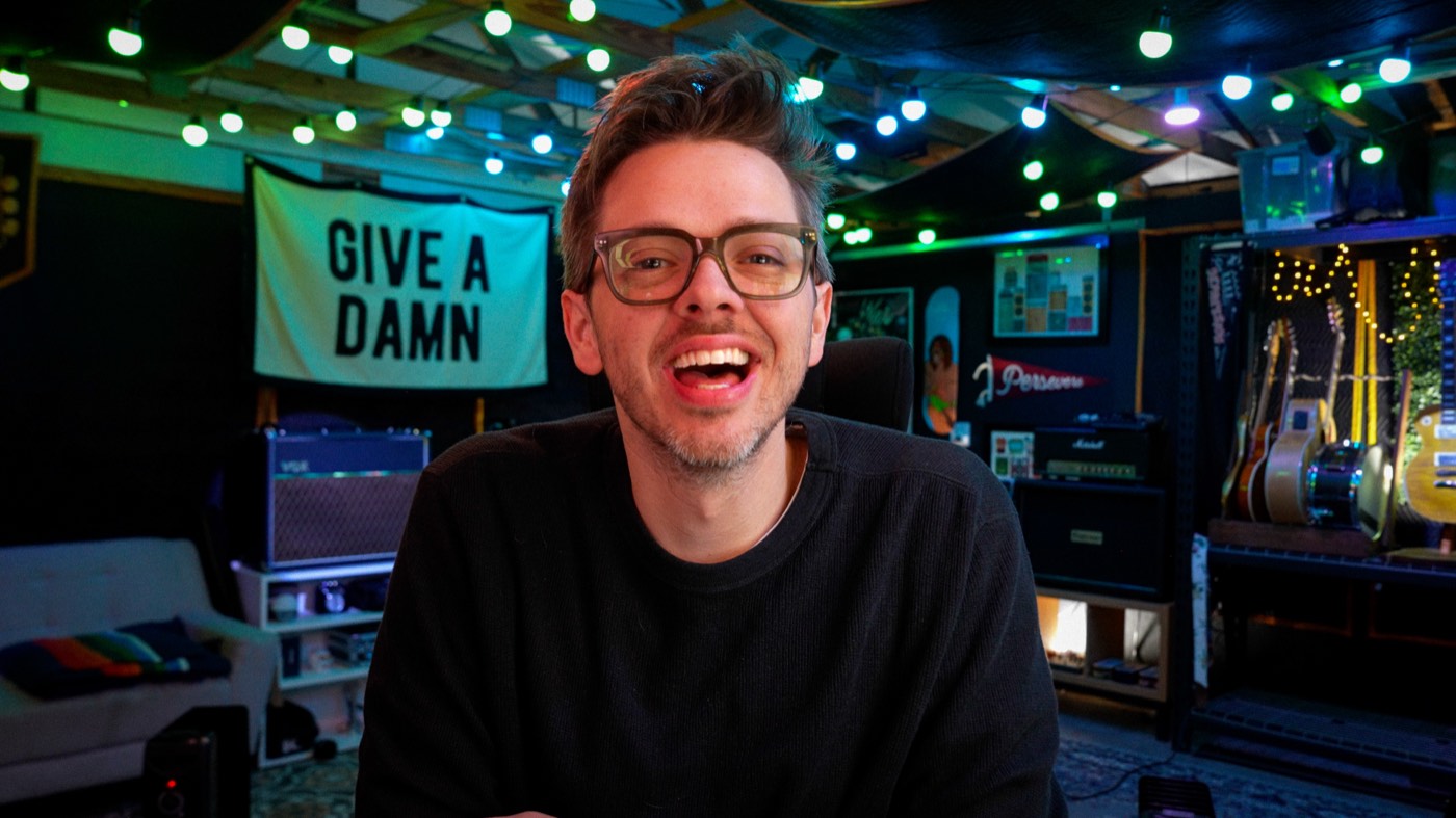 Cam laughing in his studio, the GIVE A DAMN banner and a wall of amps and guitars behind him.